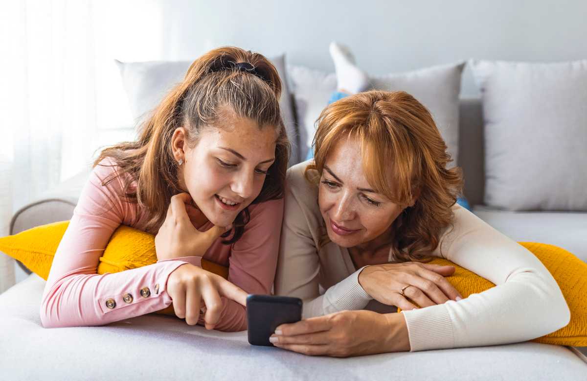 A mother and daughter laying on a bed, sharing a cell phone and talking about what is on the screen