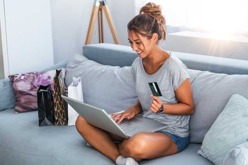 A young girl on the couch looking at her computer and holding a debit card