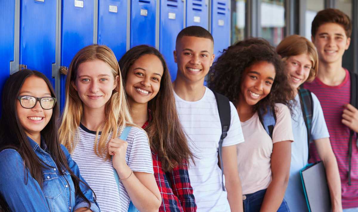 Seven teens standing next to each other, in front of their blue lockers, smiling at the camera.
